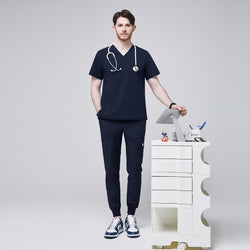 Image of Male healthcare professional wearing a navy V-neck, three-pocket scrub top and matching jogger pants with stethoscope around neck, standing beside a medical cart with supplies,Navy