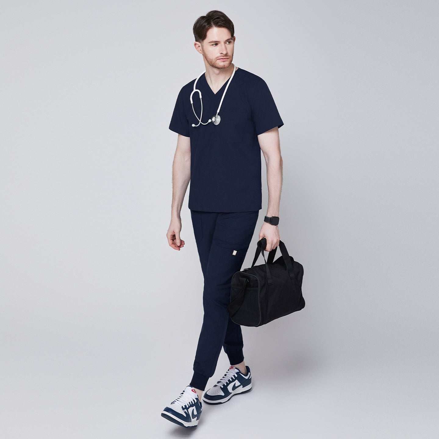 Male healthcare professional wearing navy scrub top and jogger pants, carrying a black duffel bag with stethoscope around neck, looking to the side,Navy