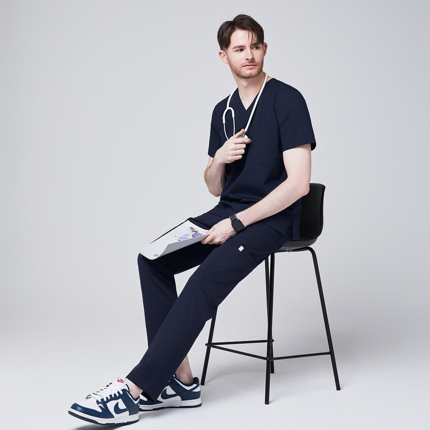 Male healthcare professional seated on a stool wearing navy scrub top and pants, holding a clipboard with stethoscope around neck, looking to the side,Navy