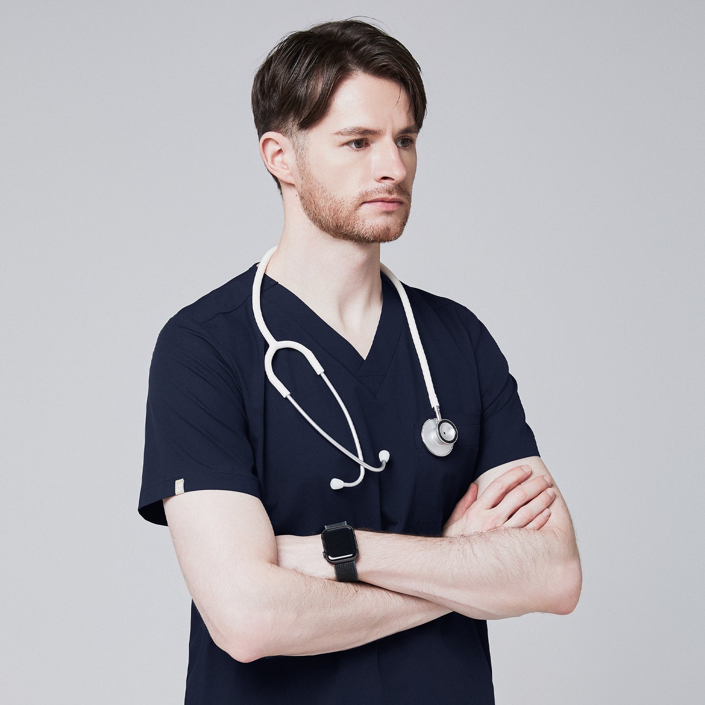 Male healthcare professional in navy V-neck scrub top with stethoscope around neck, arms crossed, looking to the side against a plain background,Navy