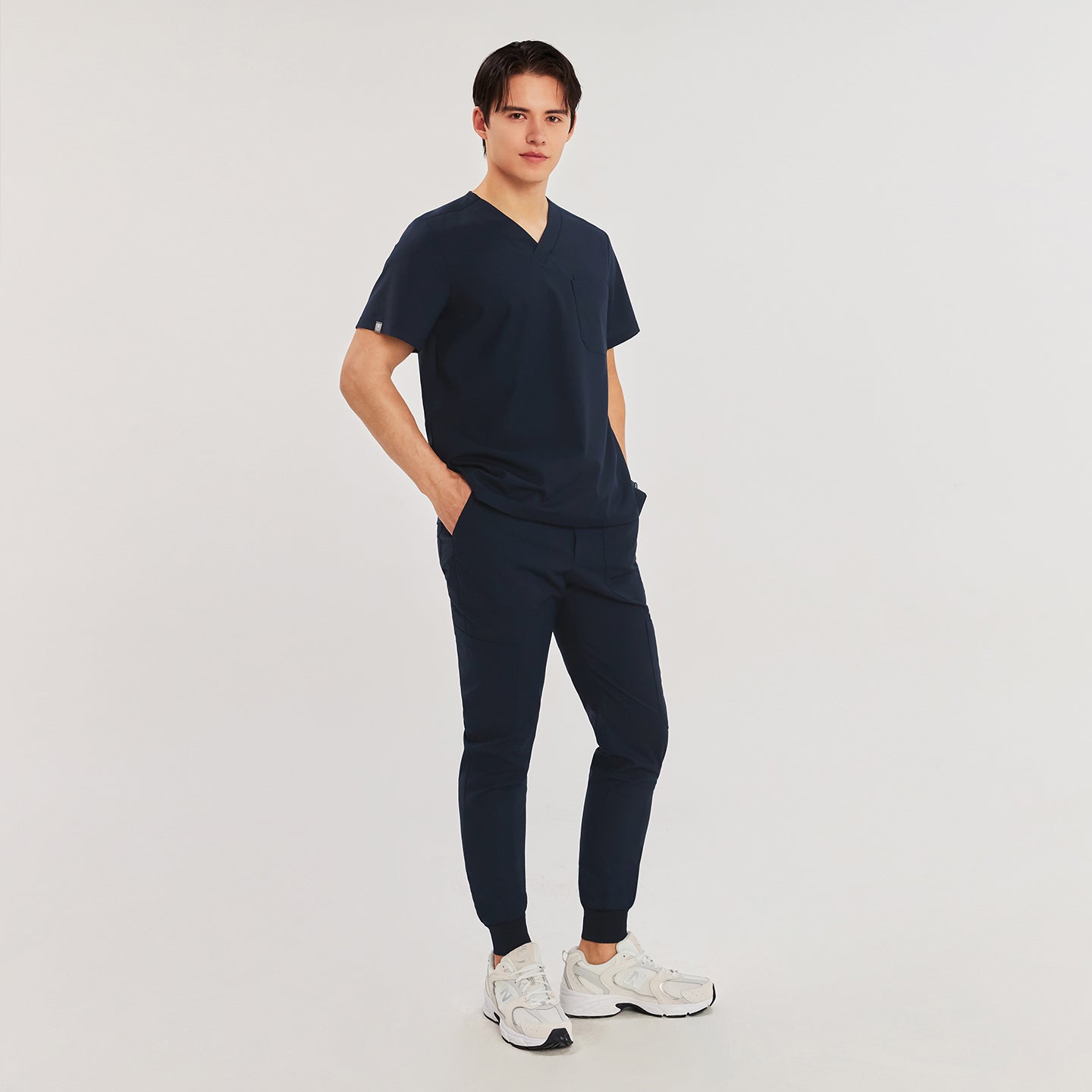  Full-body image of a man wearing jogger-style scrub pants with elastic cuffs and a V-neck scrub top, paired with white New Balance sneakers,Navy