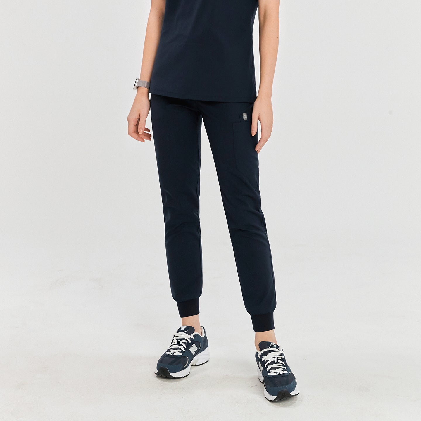 Woman wearing navy jogger scrub pants with a drawstring waist, side pockets, and ribbed cuffs, paired with a matching scrub top and navy sneakers,Navy