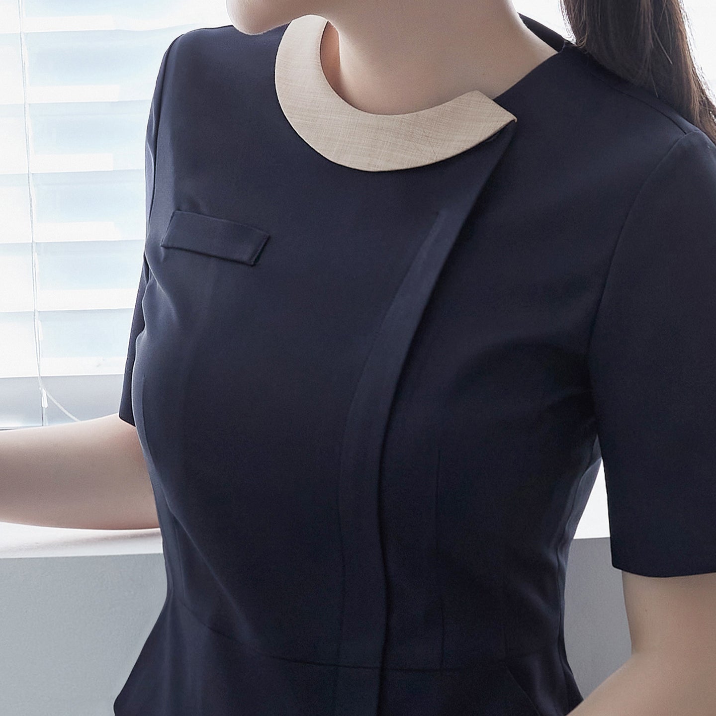  Detail view of a woman wearing a navy round-neck front zipper top with a contrasting beige collar, standing by a window,Navy