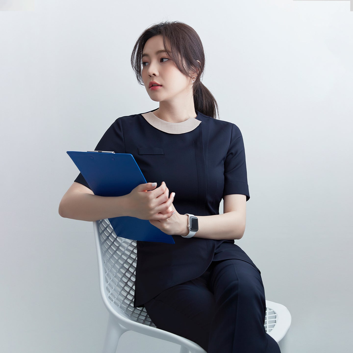  A woman in a navy round-neck front zipper top and matching pants, sitting on a white chair, holding a blue clipboard. She wears a smartwatch,Navy