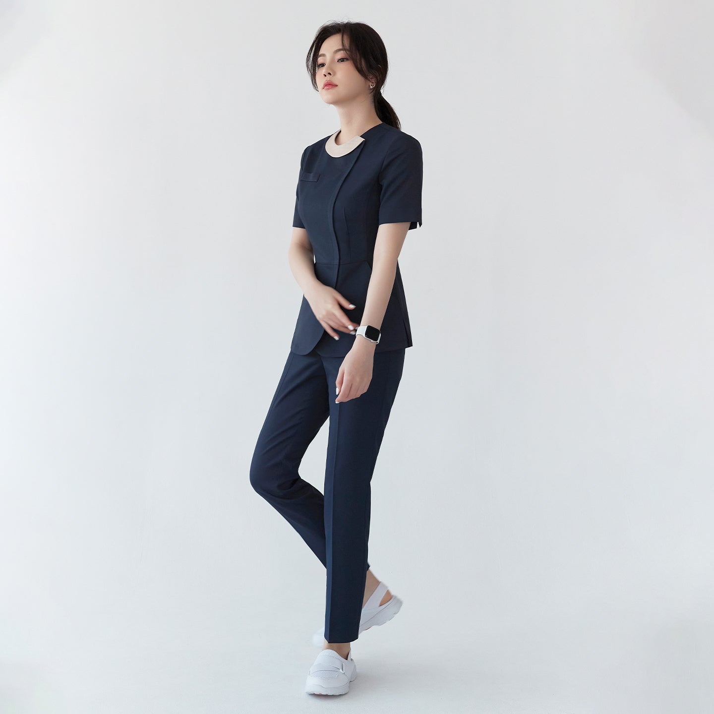 A woman in a navy round-neck front zipper top and matching pants, standing with a hand on her stomach. She wears white shoes and a smartwatch,Navy