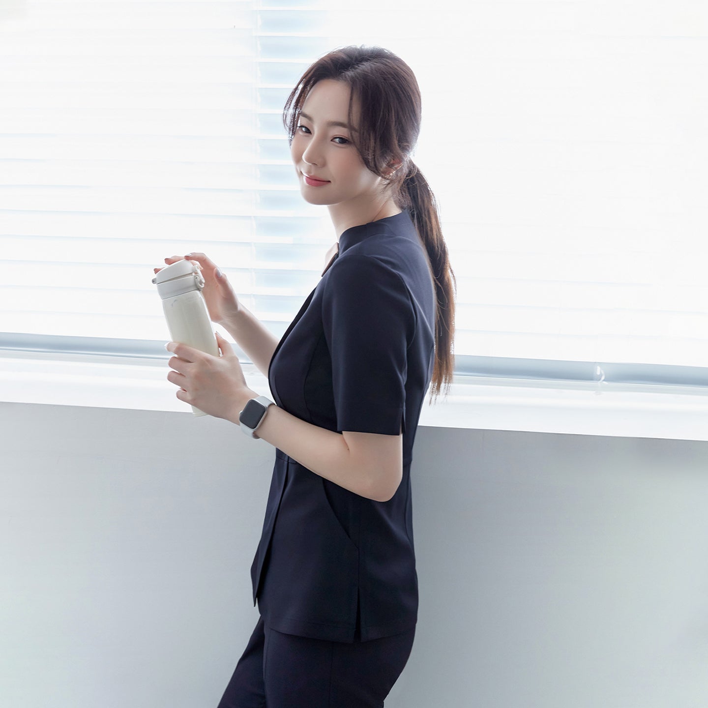 A woman in a navy round-neck front zipper top and matching pants, standing by a window, holding a white tumbler, and wearing a smartwatch,Navy
