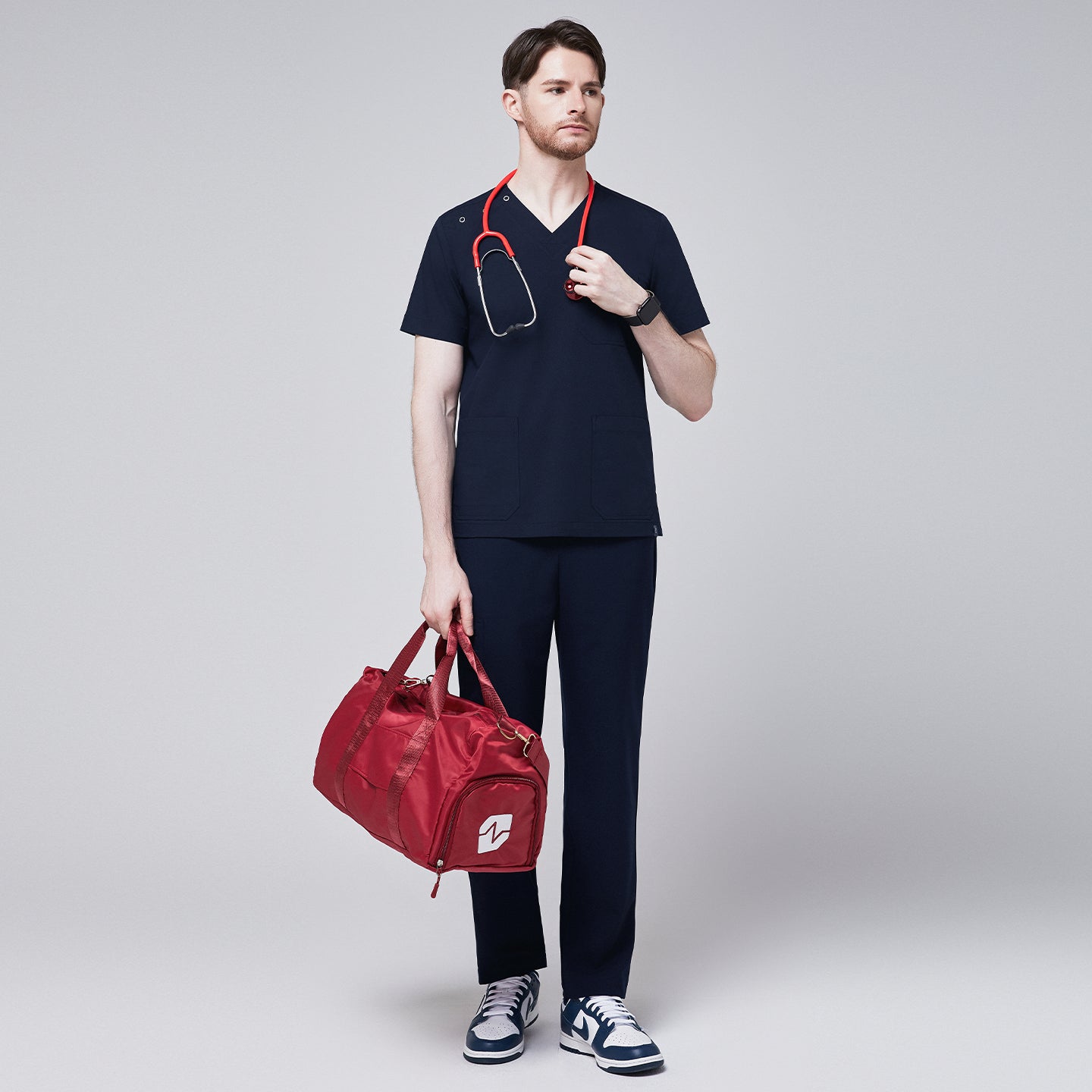 Full-body view of a man wearing a navy scrub top with button details and matching pants, holding a red duffle bag and a stethoscope, exuding a professional and functional look,Eco Navy