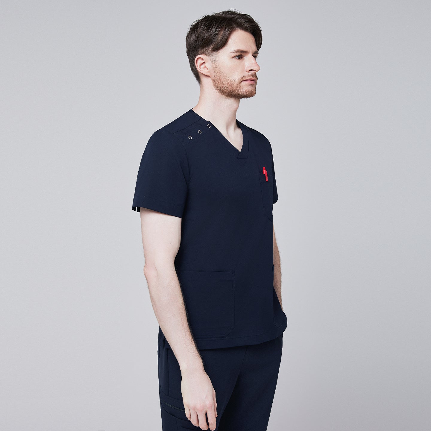 Side angle of a man wearing a navy scrub top with shoulder button detailing, featuring a red pen in the chest pocket and hands casually resting in the front pockets,Eco Navy