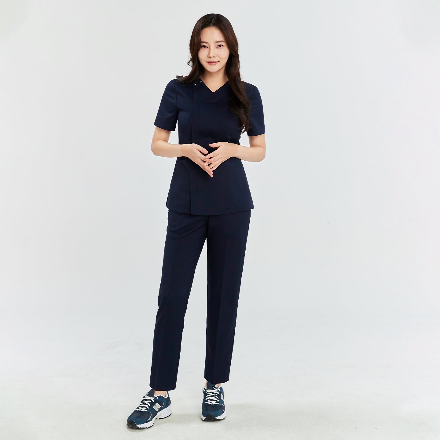 Woman in a navy front zipper scrub top and straight-leg pants, standing with hands clasped, wearing navy sneakers,Navy