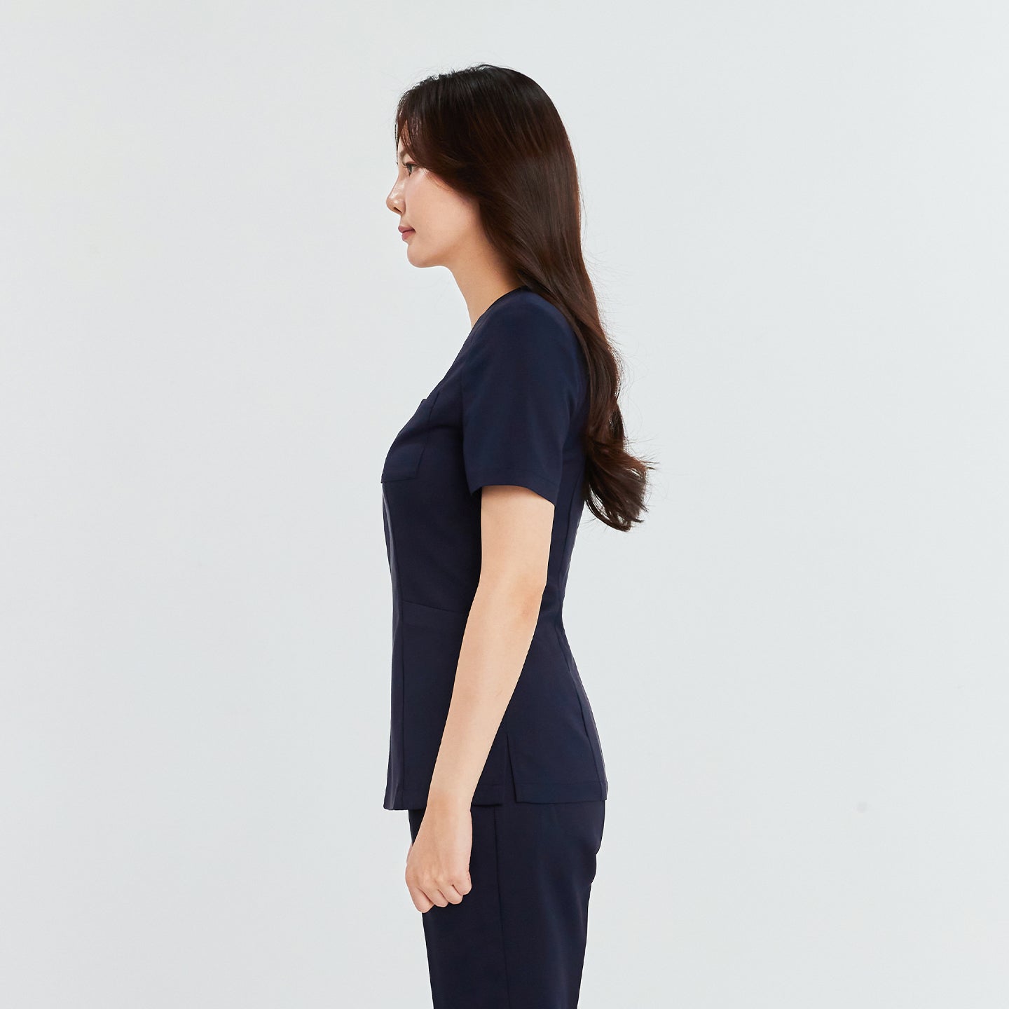 Woman in a navy front zipper scrub top with patch pockets and matching straight-leg pants, standing in profile view,Navy