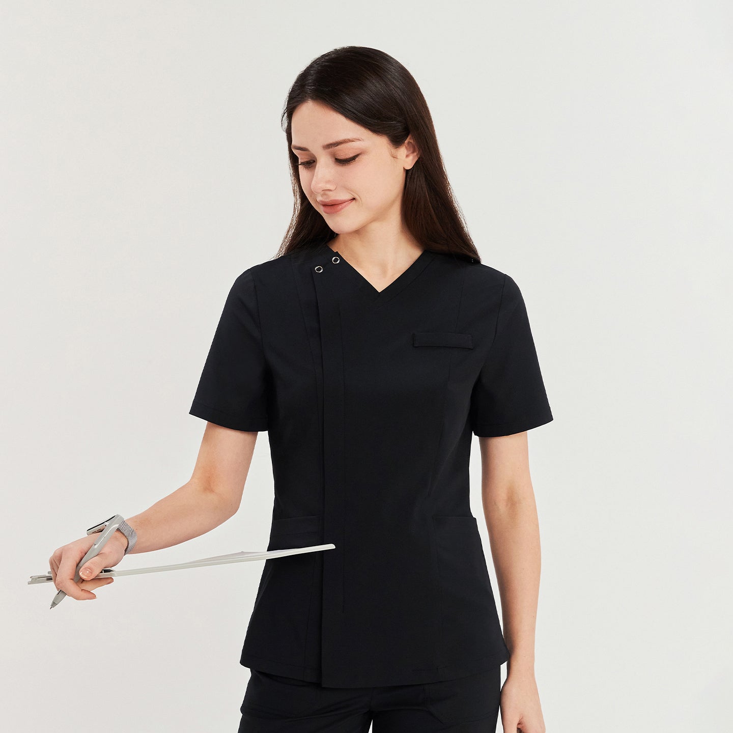 Woman in a black front zipper scrub top with patch pockets and matching straight-leg pants, holding a clipboard, looking down,Black