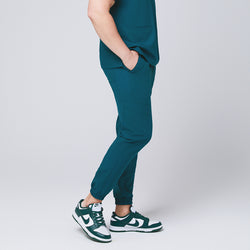 Image of Ocean teal jogger scrub pants made of stretchy fabric and matching ocean teal scrub top, with teal sneakers and hands in pocket,Ocean teal