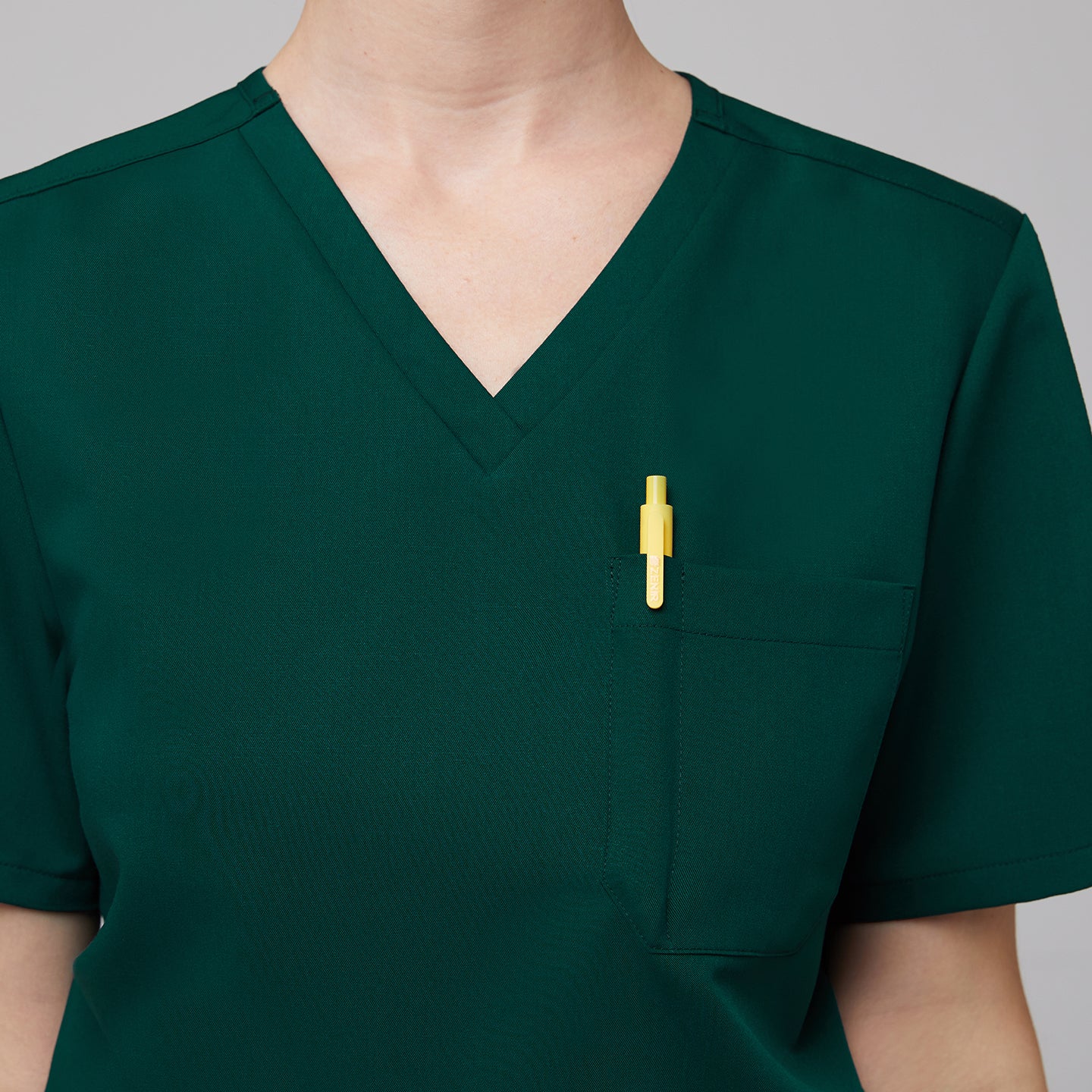 Close-up of a pine green scrub top featuring a V-neck design and a chest pocket holding a yellow pen,Pine Green