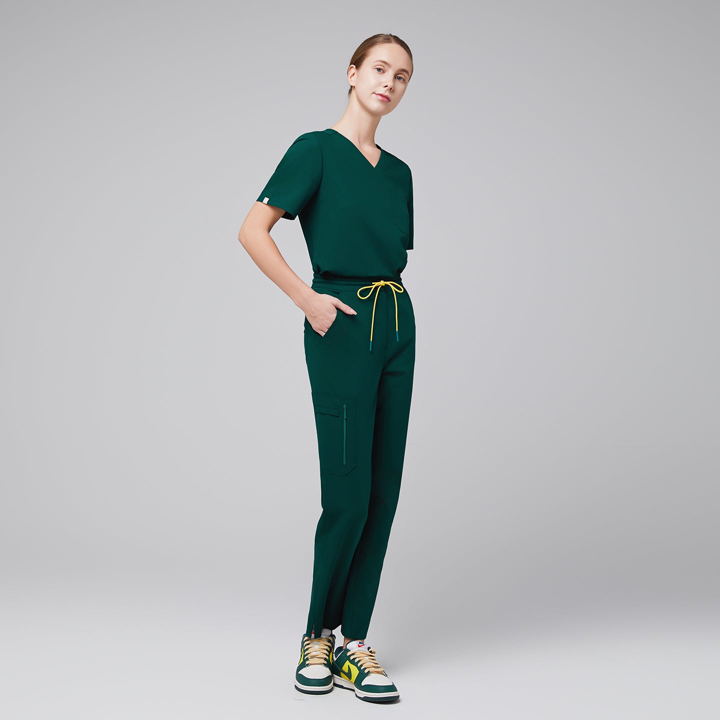 A woman dressed in a pine green scrub set, featuring a relaxed V-neck top with three pockets and tapered pants with yellow drawstring accents, styled with sporty sneakers,Pine Green