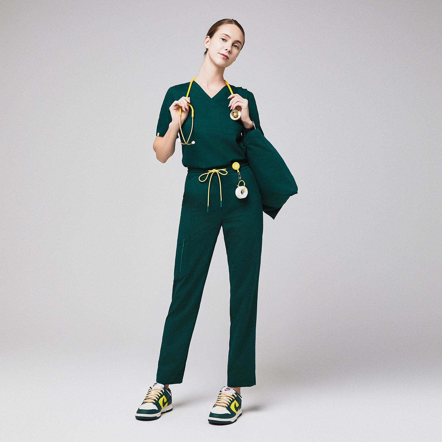 Female healthcare professional in green scrubs with zipper pocket pants and yellow drawstring, holding a stethoscope and jacket,Pine Green