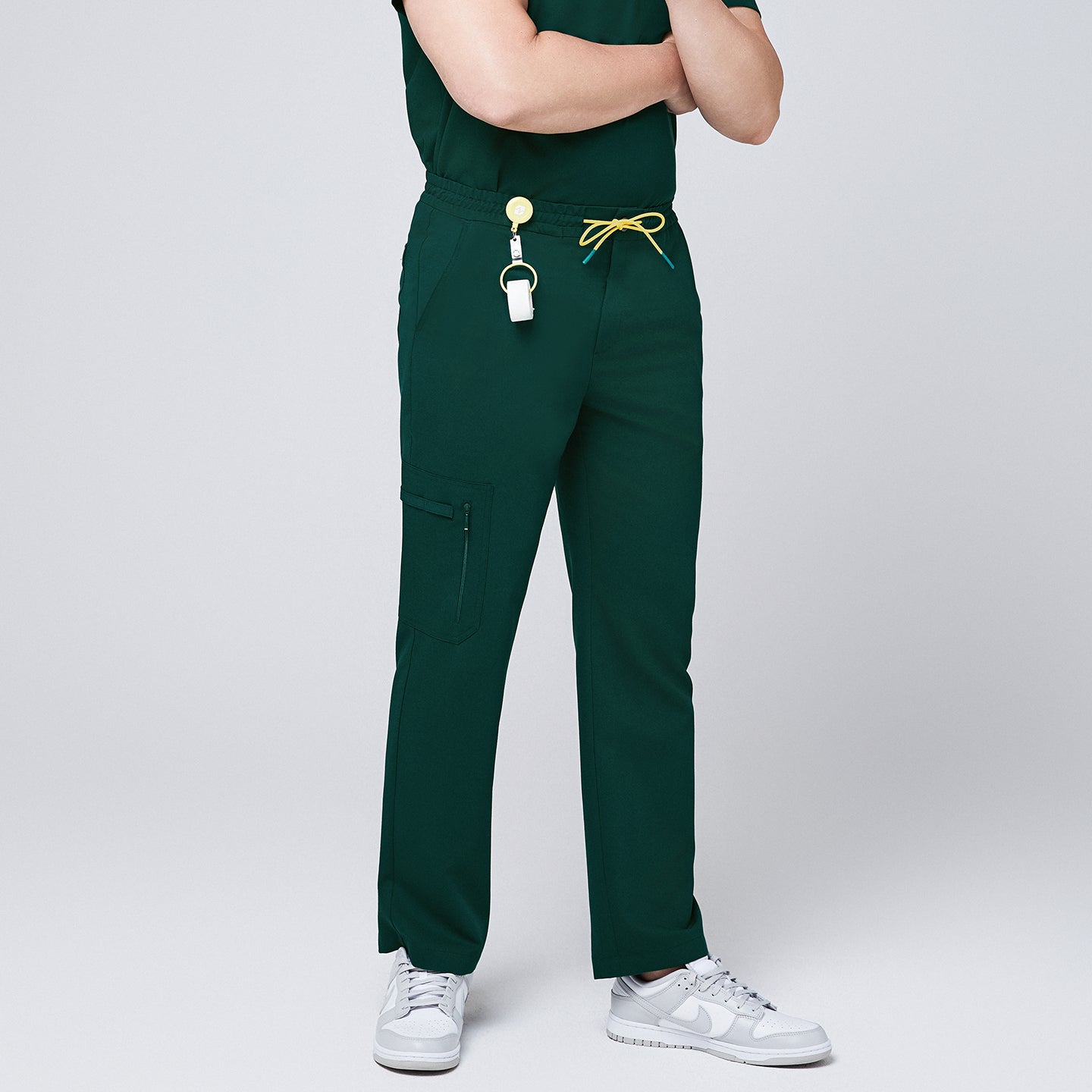 Male model wearing pine green straight-leg scrub pants with zipper pockets and a yellow drawstring, accessorized with a badge reel and white sneakers,Pine Green