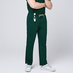 Image of Male model wearing pine green straight-leg scrub pants with zipper pockets and a yellow drawstring, accessorized with a badge reel and white sneakers,Pine Green