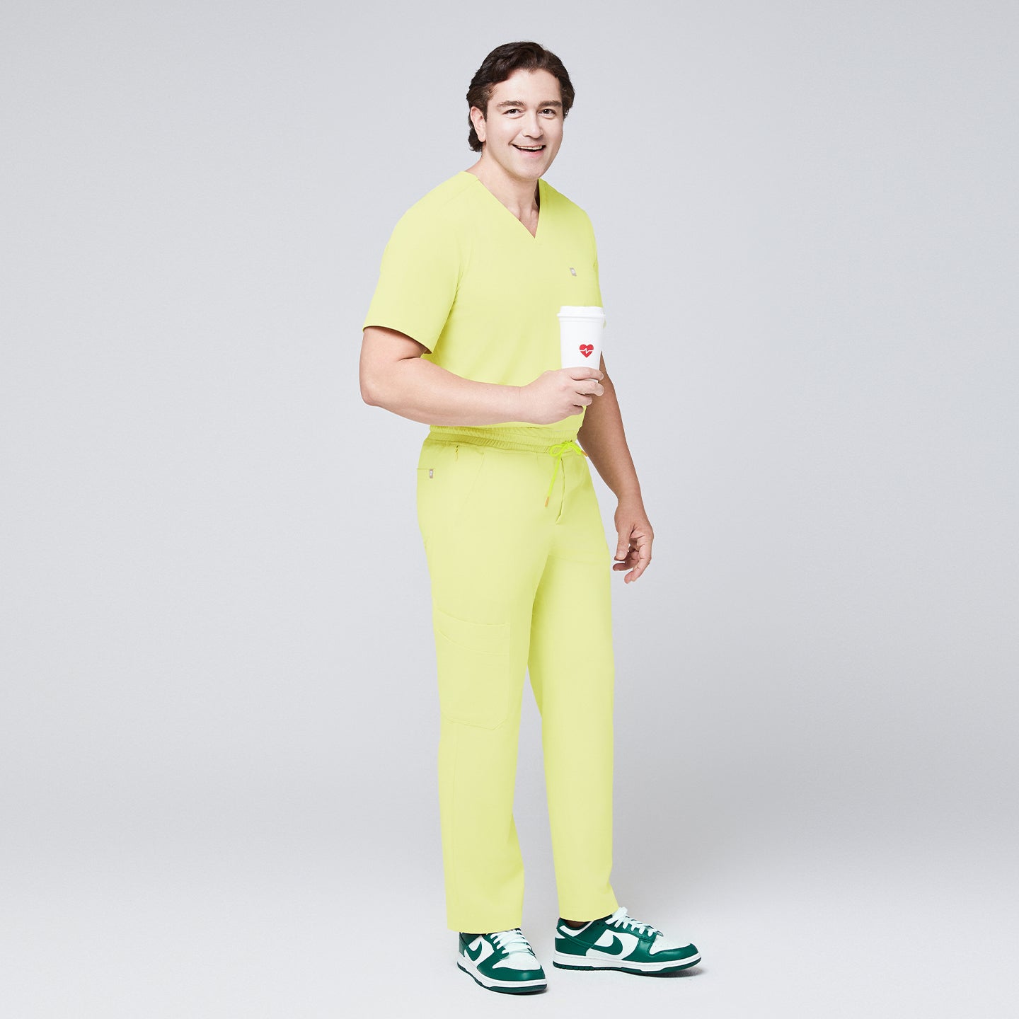 Man wearing pineapple yellow straight-leg scrub pants with a drawstring waistband and matching V-neck top, holding a coffee cup and styled with green sneakers,Pineapple Yellow