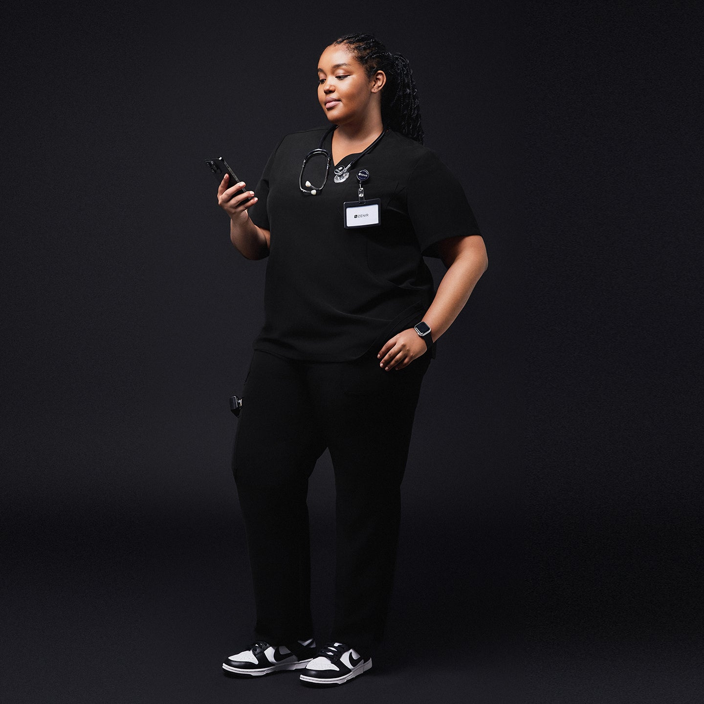 Medical professional in black scrubs holding a smartphone, with a stethoscope around her neck and a name tag, standing confidently against a dark background,Rich Black