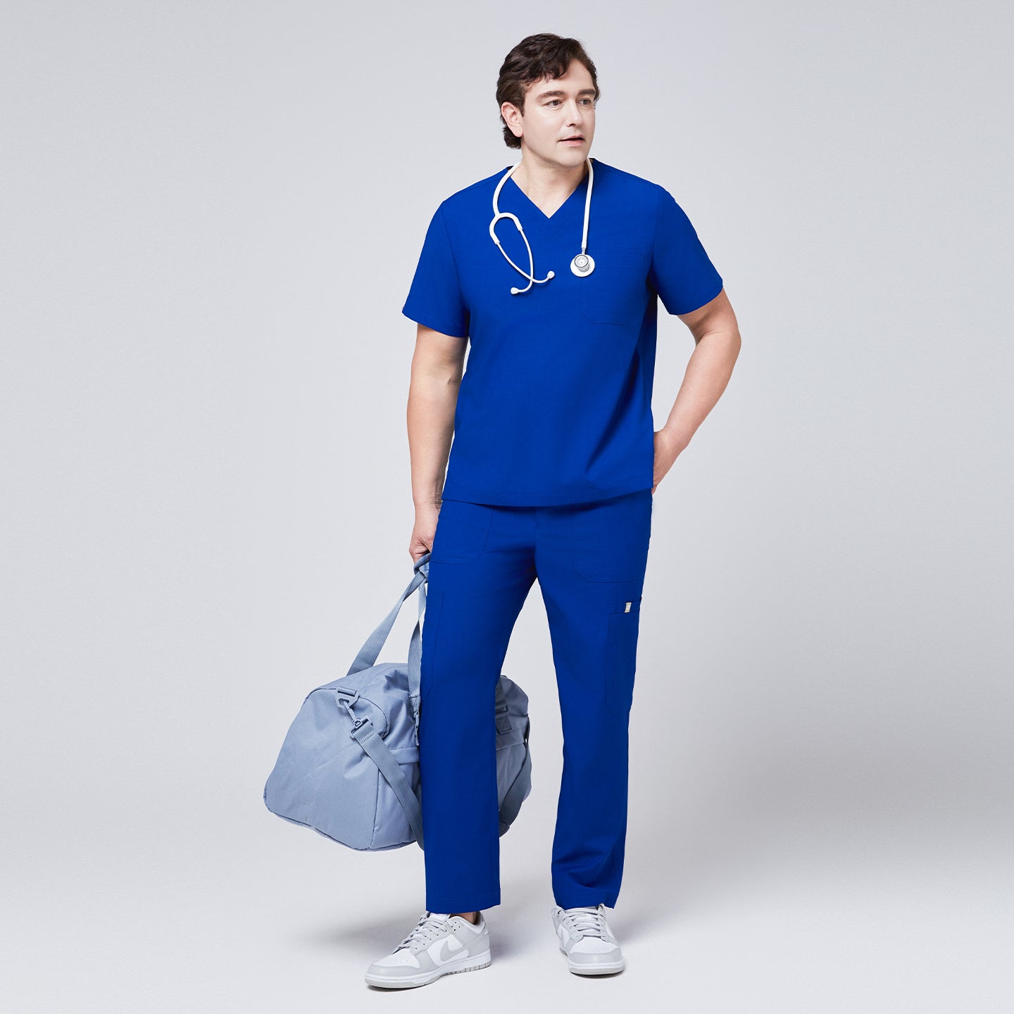 Man dressed in a royal blue V-neck scrub top and matching pants, holding a light blue duffel bag and wearing a stethoscop,Royal Blue