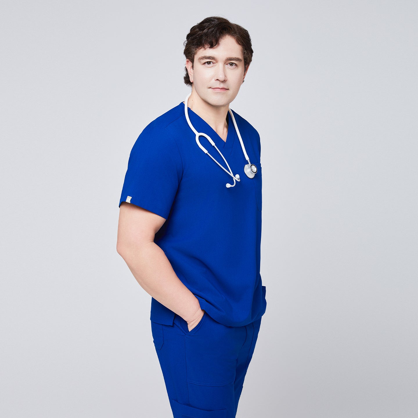 Man in a royal blue V-neck scrub top with a white stethoscope around his neck, paired with matching pants, hands in pockets,Royal Blue