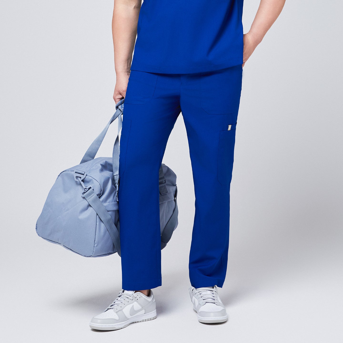 Close-up of royal blue 8-pocket scrub pants paired with a matching scrub top, white sneakers, and a light blue duffle bag,Royal Blue