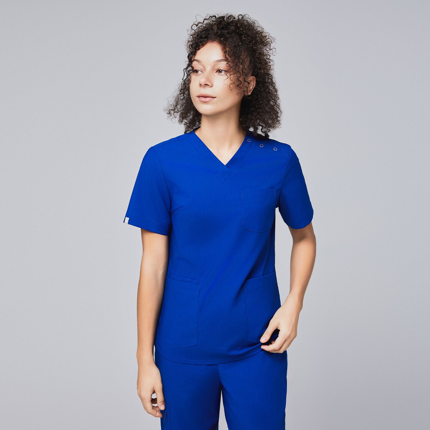 A woman wearing a royal blue scrub top with shoulder button details and matching scrub pants, looking to the side with a confident expression,Royal Blue