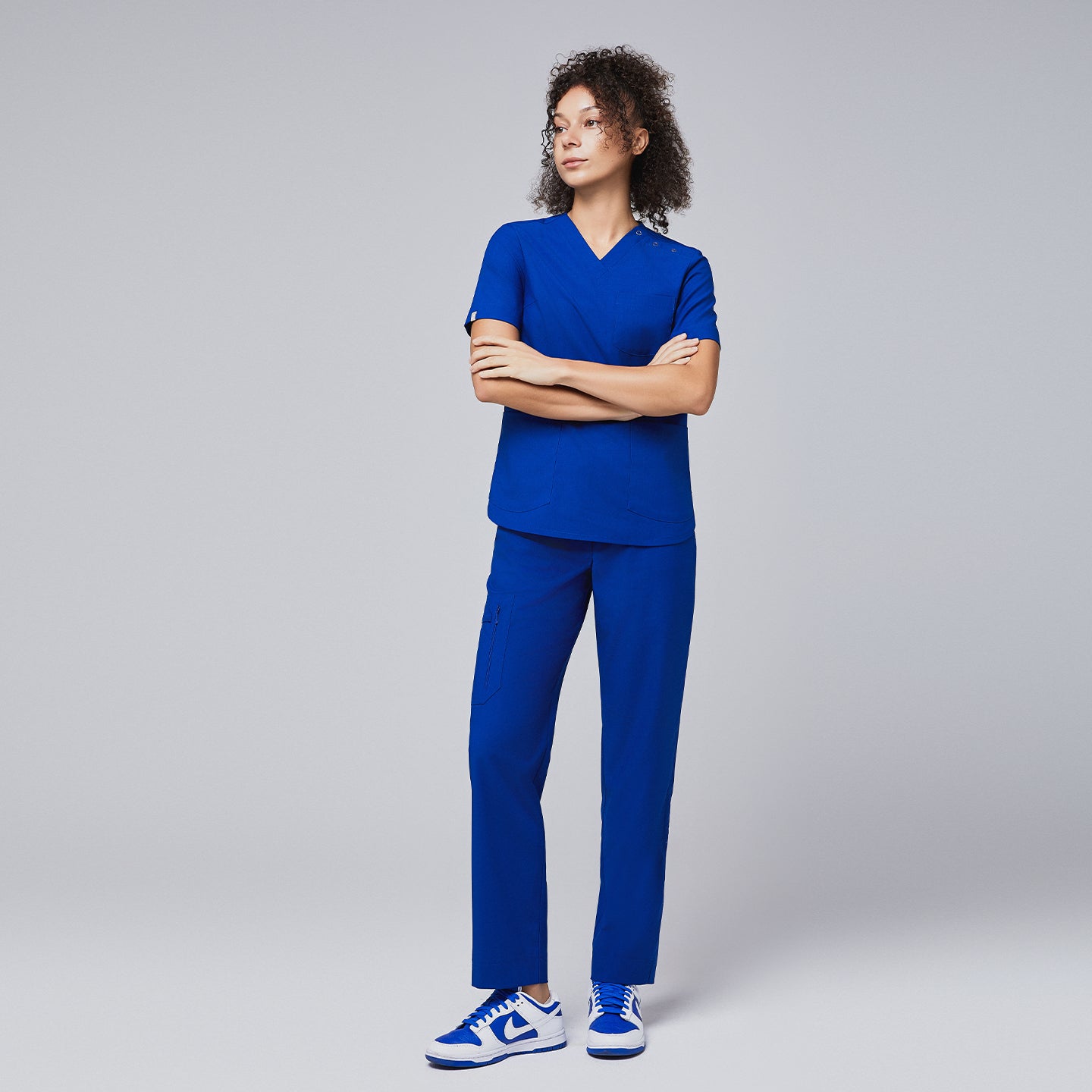 A woman wearing a royal blue scrub set with a V-neck top featuring shoulder button details and straight-leg pants, standing confidently with arms crossed,Royal Blue