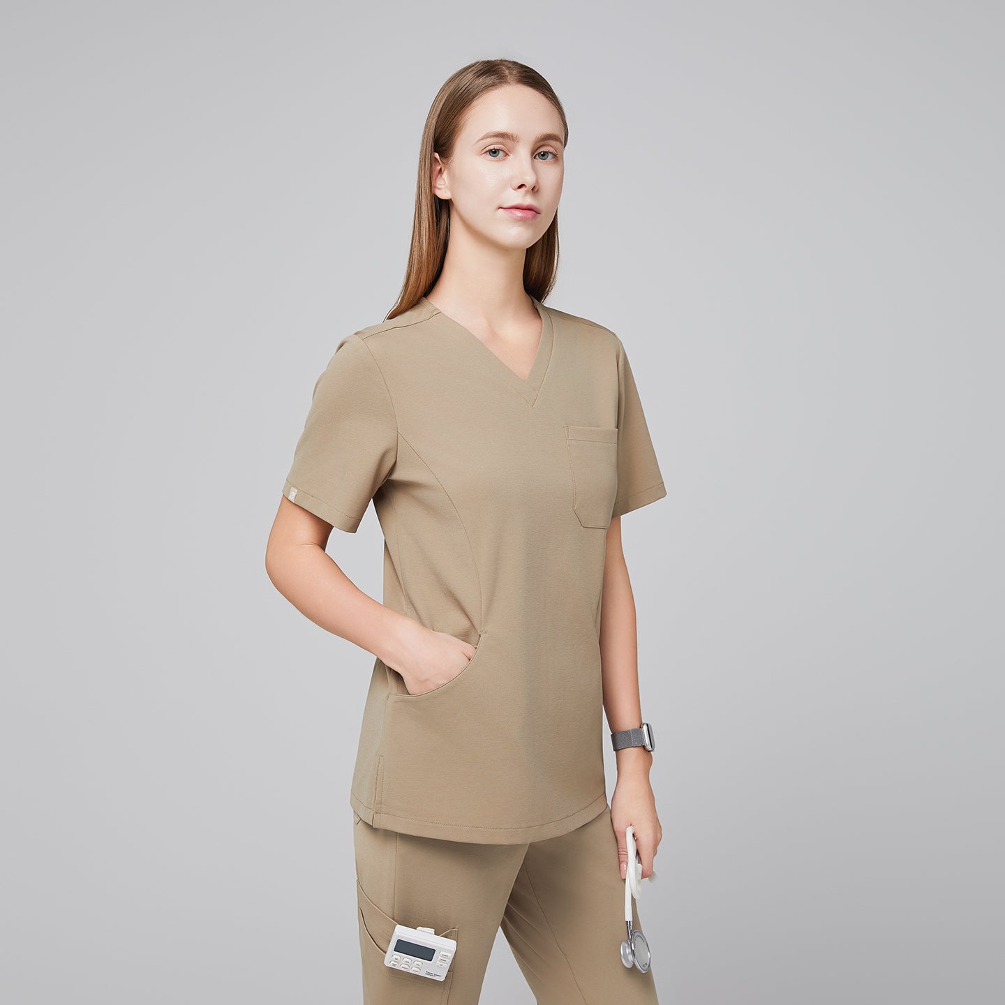 Female model in sand beige soft stretch scrub top with hands in front pockets, shown in a half-side view, holding a stethoscope,Sand Beige