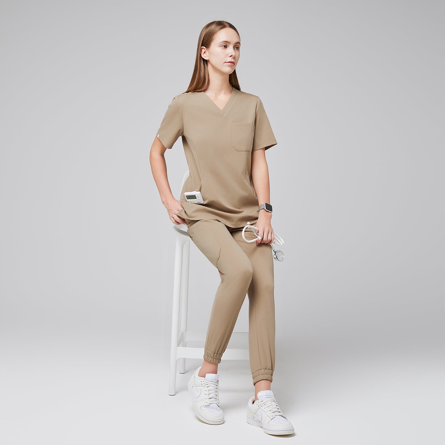 Professional scrub set styled with a relaxed fit top and elastic ankle jogger pants, paired with white sneakers, modeled in a seated position on a stool for a polished and comfortable appearance,Sand Beige