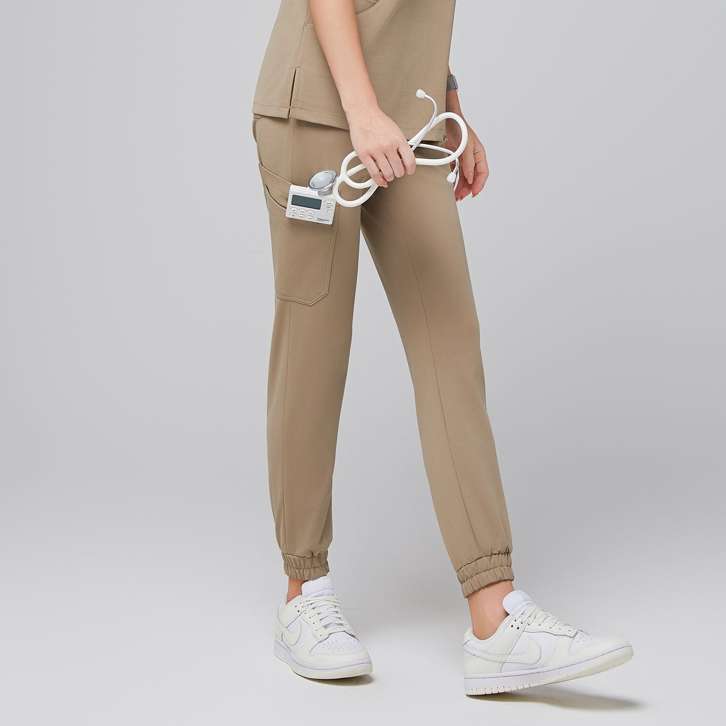 Female wearing sand beige jogger scrub pants with side pockets, holding a stethoscope, paired with white sneakers,Sand Beige