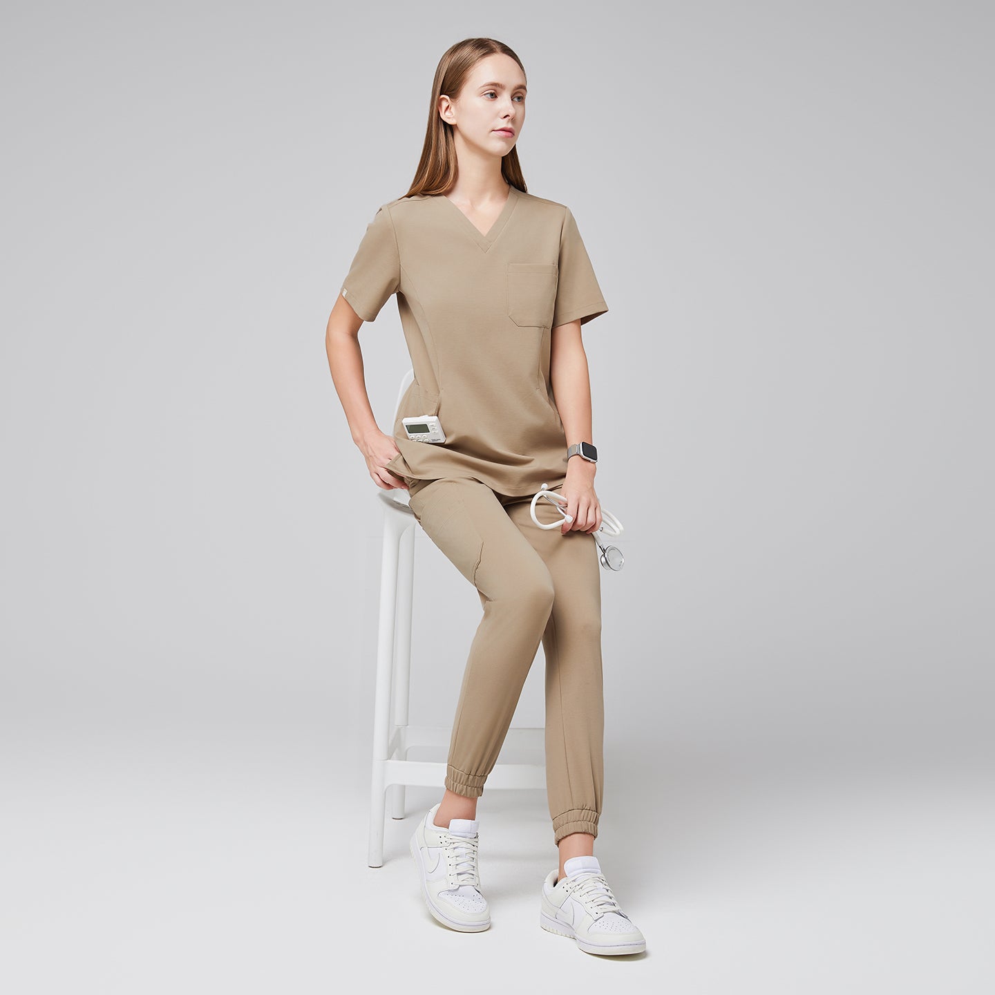 Female wearing sand beige scrub top and jogger pants, sitting on a white stool, holding a stethoscope, paired with white sneakers,Sand Beige