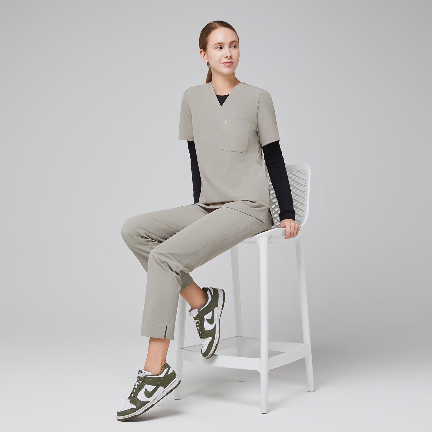 Female healthcare professional sitting on a white chair, wearing a sand khaki scrub set with a black undershirt and green sneakers,Sand Khaki