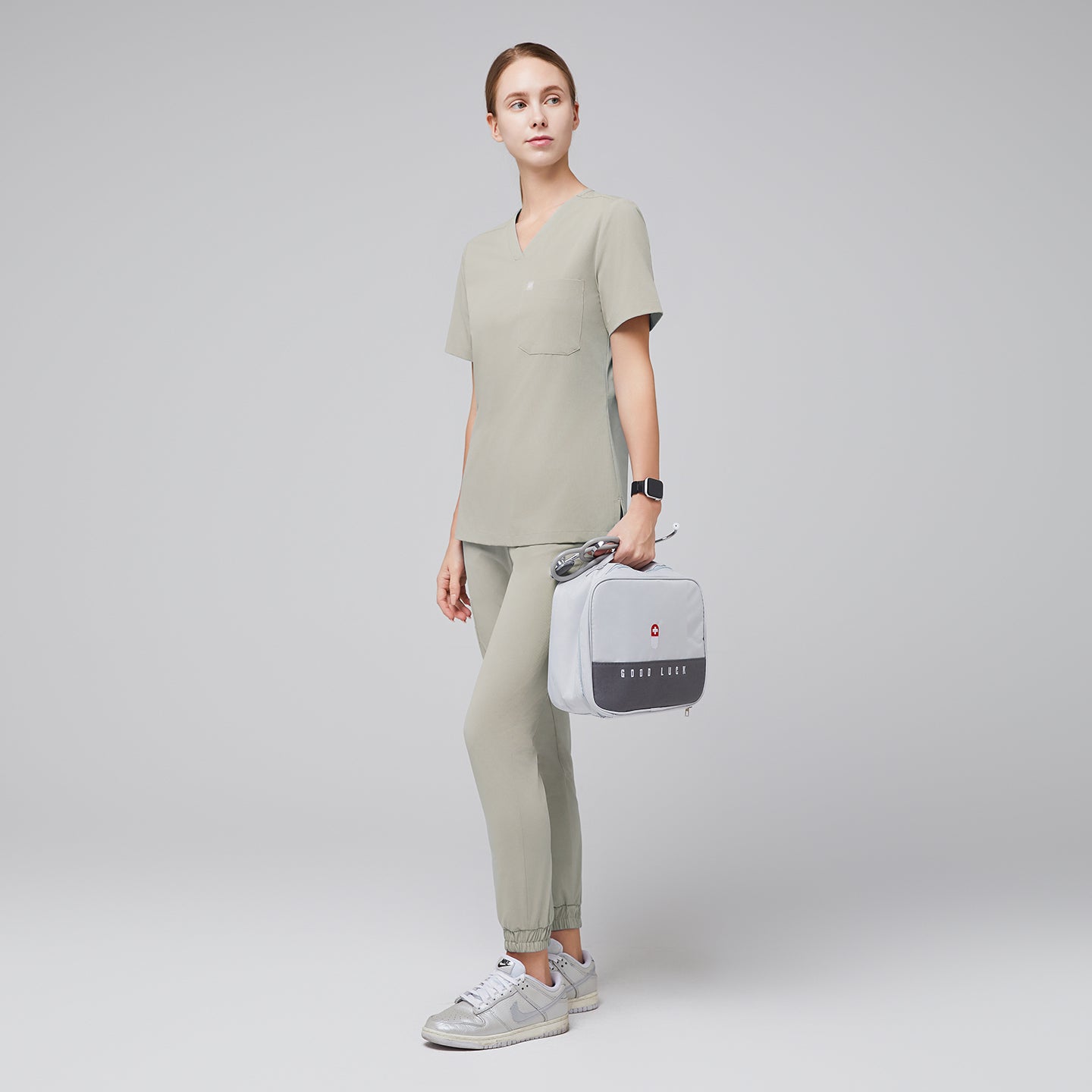 Full-body side view of a model wearing sand khaki jogger scrub pants paired with a matching V-neck scrub top. The outfit is styled with silver sneakers and accessorized with a gray medical pouch, creating a modern and professional look, Sand Khaki