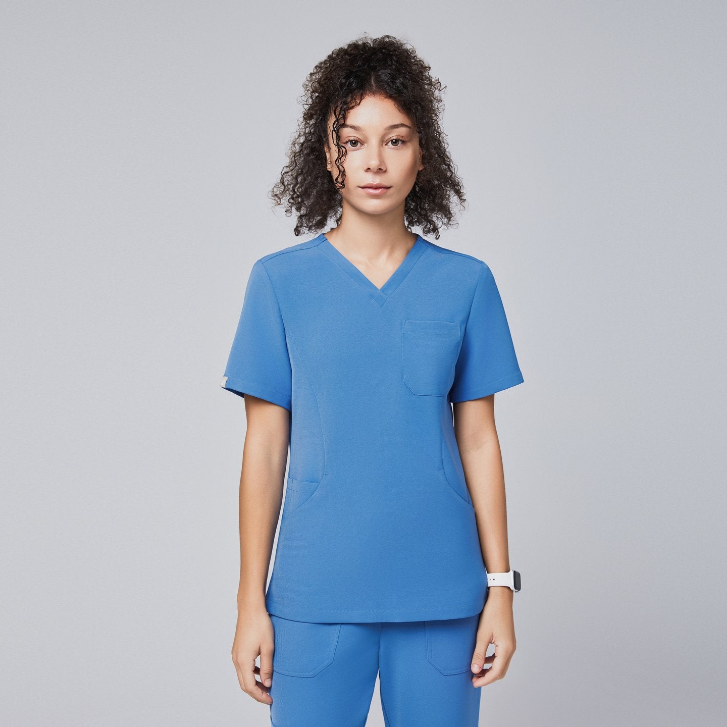 Model with curly hair wearing the sky blue scrub top, featuring a V-neck, short sleeves, and a chest pocket against a neutral background,Sky Blue