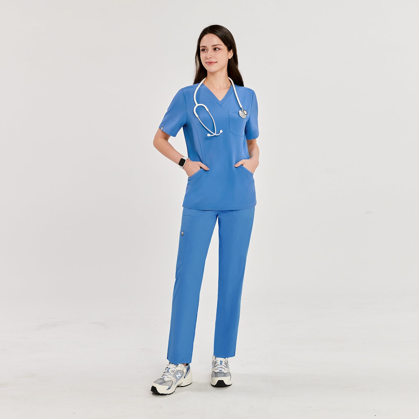 Woman wearing a sky blue scrub top and matching pants, with a stethoscope around her neck, hands in pockets, standing and looking to the side,Sky Blue