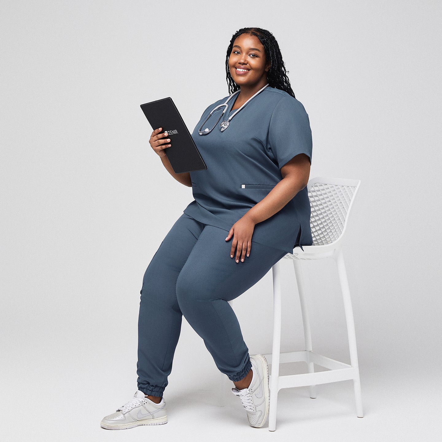 Plus-size healthcare worker in smoky blue scrub set with stethoscope holding a Zenir clipboard while seated,Smoky Blue