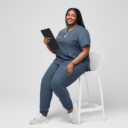 Plus-size healthcare worker in smoky blue scrub set with stethoscope holding a Zenir clipboard while seated,Smoky Blue