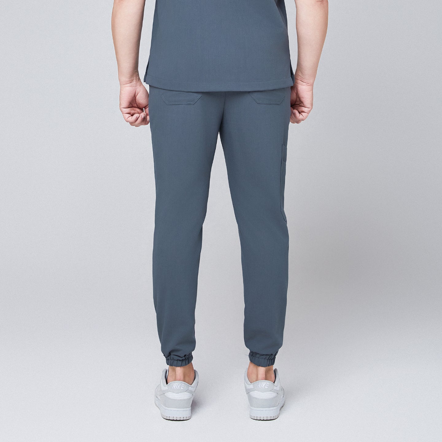 Back view of smoky blue jogger scrub pants with elastic cuffs and flap pockets, paired with gray Nike sneakers,Smoky Blue