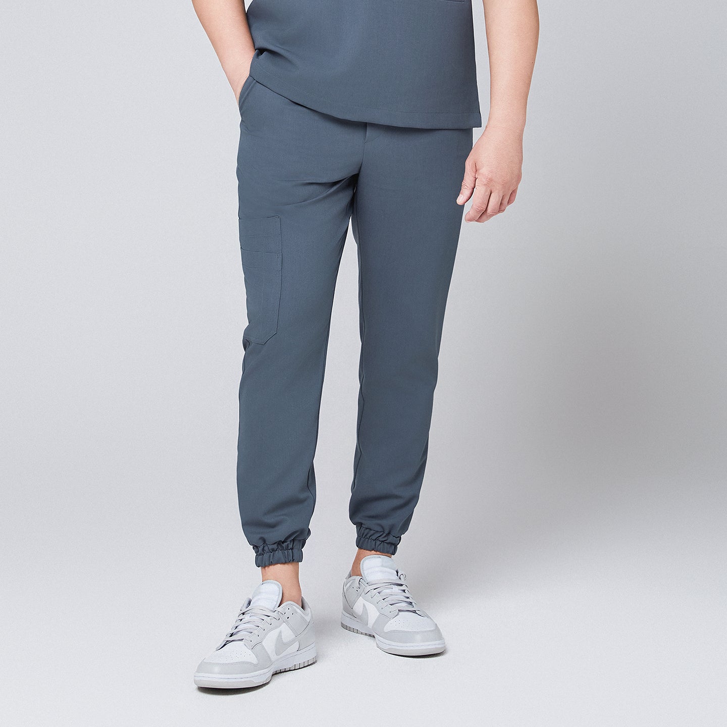 Front view of smoky blue jogger scrub pants with elastic cuffs and a side cargo pocket, paired with gray Nike sneakers,Smoky Blue