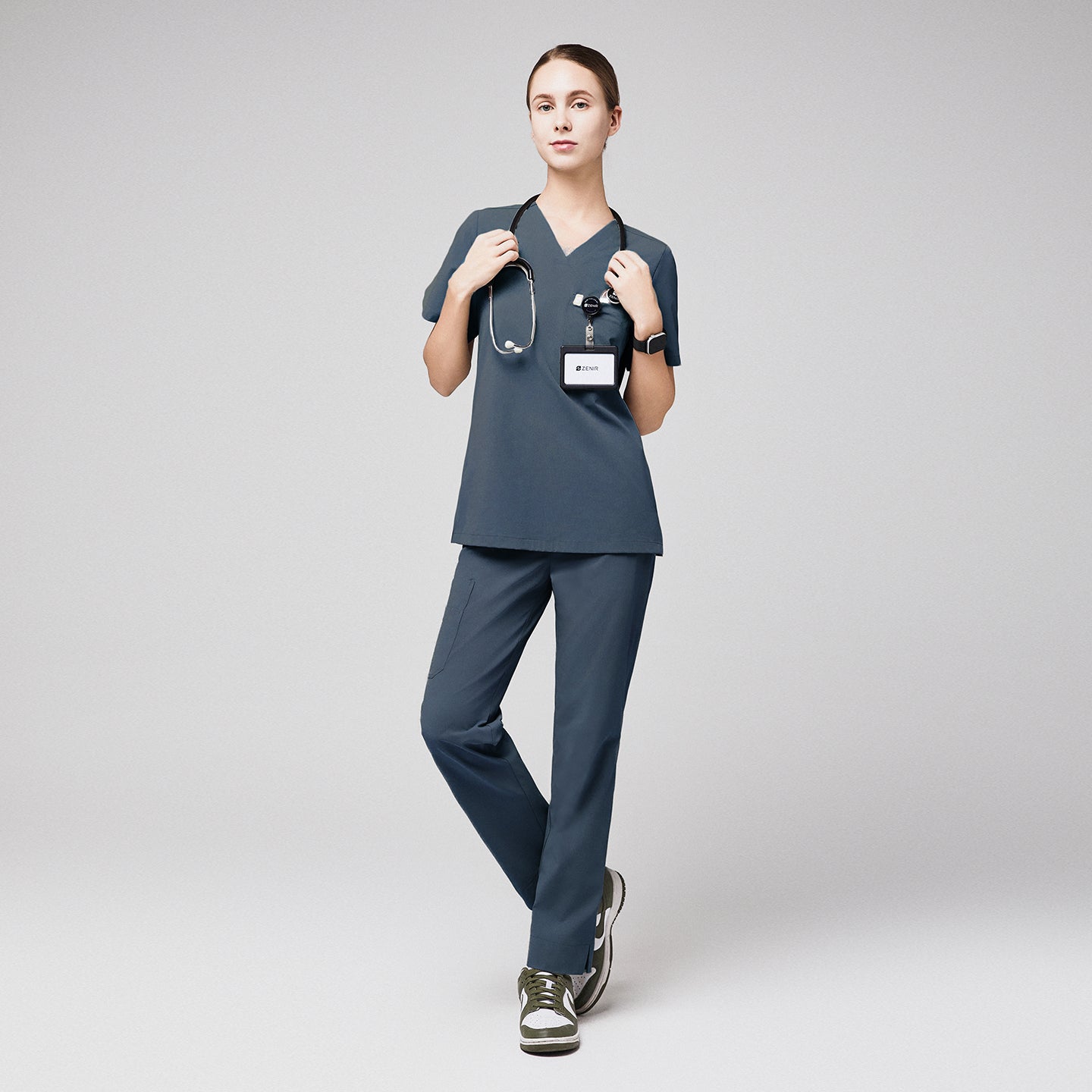 Female in smoky blue scrubs with a stethoscope and ID badge, standing with crossed legs, paired with green and white sneakers,Smoky Blue