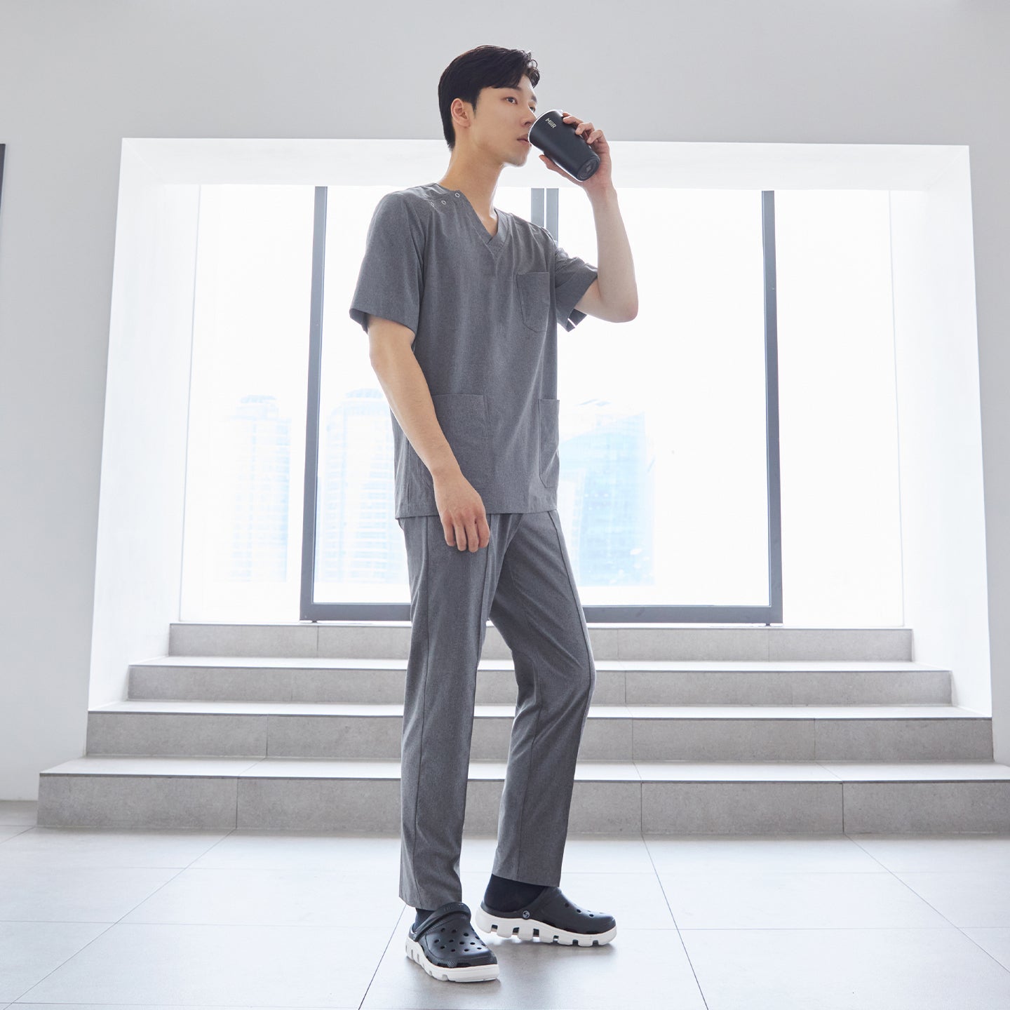 Man wearing front zipper scrub top with chest and side pockets, paired with banding scrub pants, standing indoors and drinking from a cup,Ash Gray