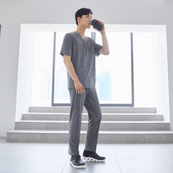 Image of Man wearing front zipper scrub top with chest and side pockets, paired with banding scrub pants, standing indoors and drinking from a cup,Ash Gray