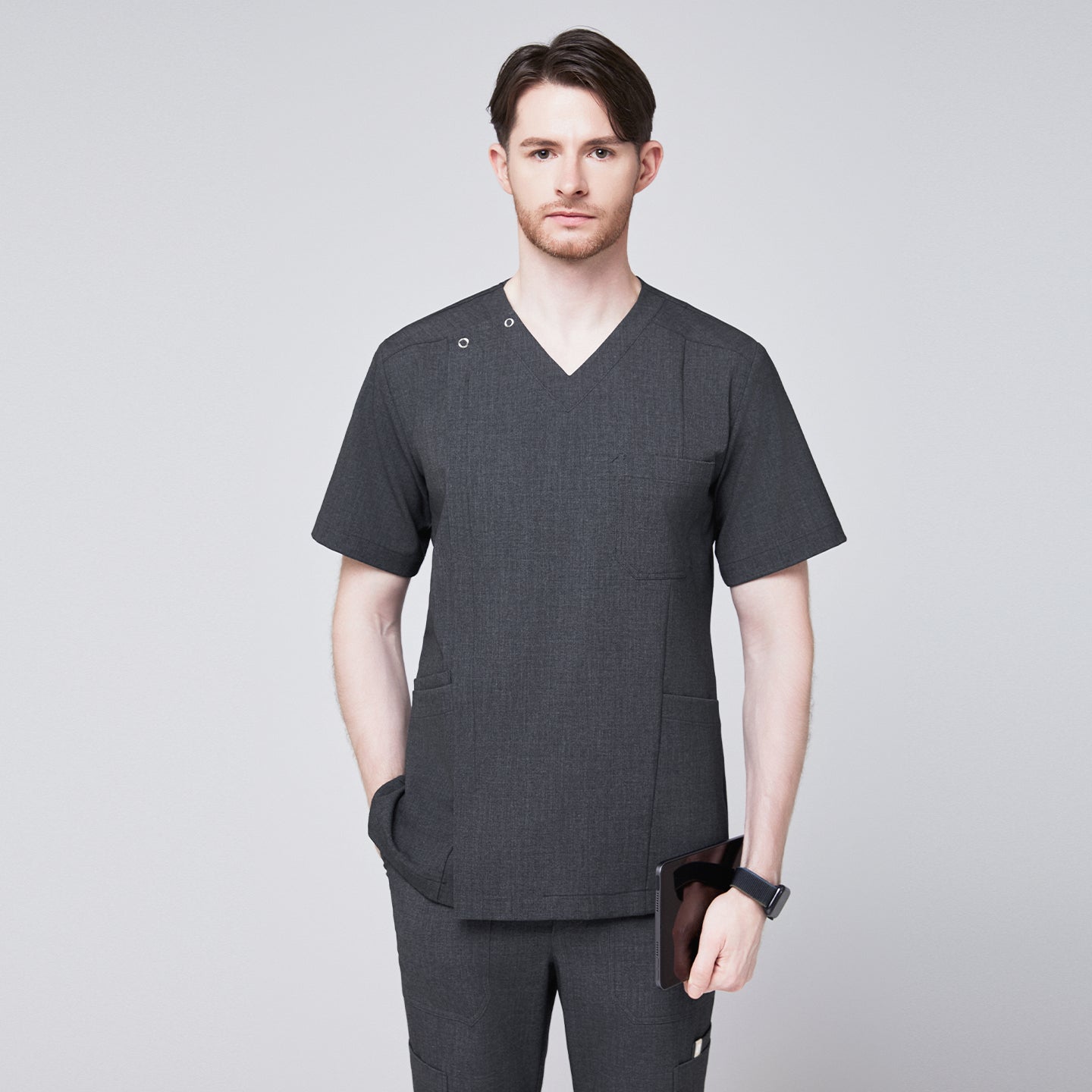 Man wearing a soft gray scrub top with a V-neck and shoulder button detail, paired with matching pants, holding a tablet and posing with one hand in his pocket,Charcoal Gray