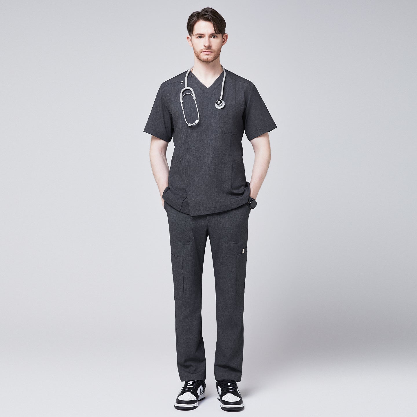 Man in a soft gray scrub top with a V-neck and shoulder button detail, styled with matching pants, wearing a stethoscope around his neck and posing with hands in pockets,Charcoal Gray