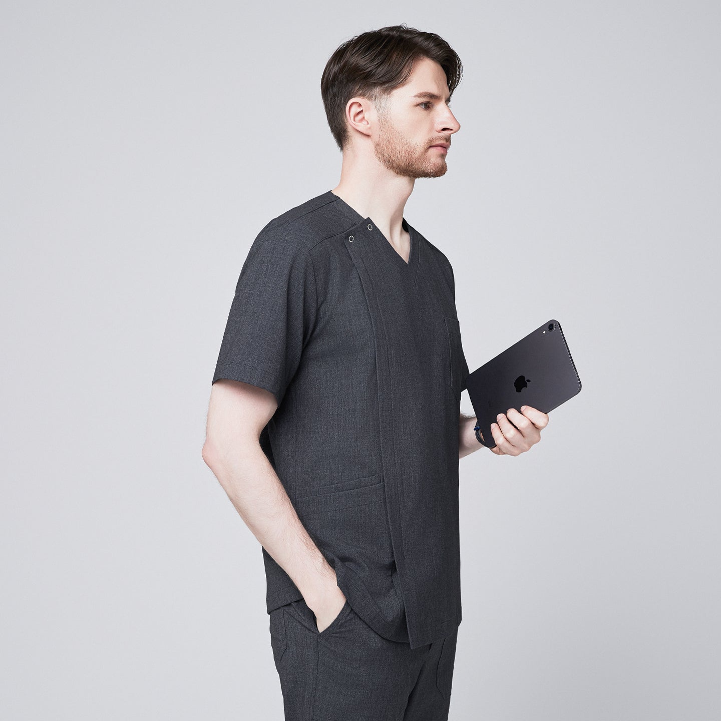 Man in a soft gray scrub top with a V-neck and shoulder button detail, standing sideways while holding a tablet and placing one hand in his pocket,Charcoal Gray