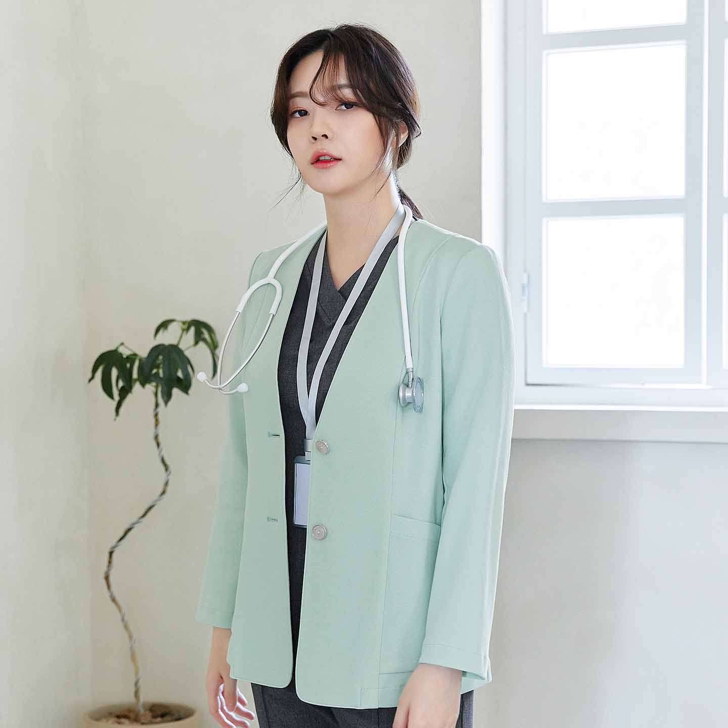 A woman wearing a stethoscope around her neck and a mint-colored jacket, standing by a window with a potted plant in the background,Soft Mint