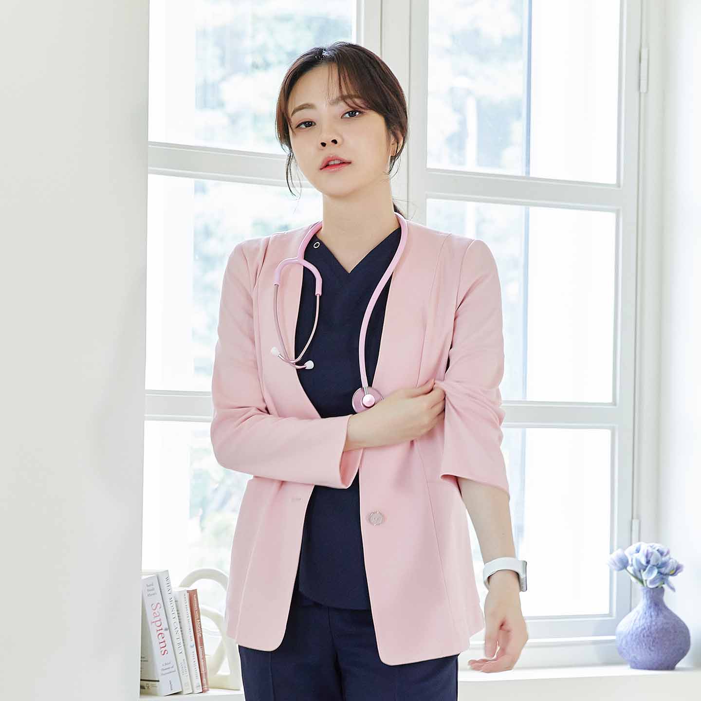 Woman in soft pink Zenir jacket over navy top, pink stethoscope, standing with arm crossed, in a room with a window and books,Soft Pink
