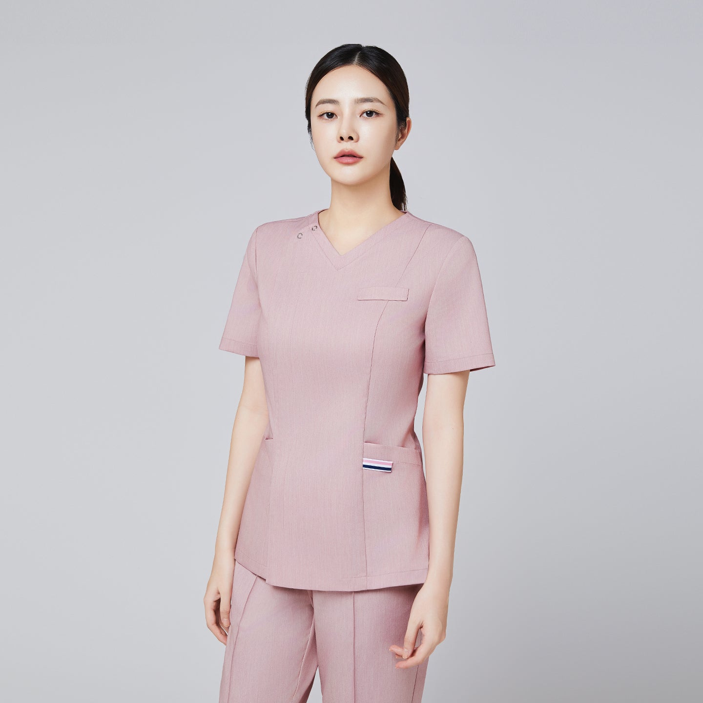 Female model wearing a soft pink scrub top with front zipper detail and matching pants, posing with a professional expression.Soft Pink