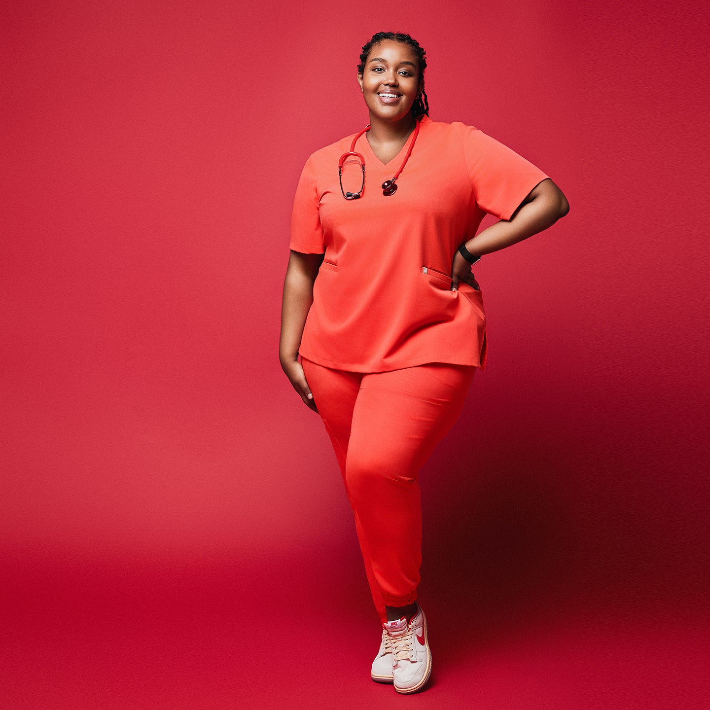 Plus-size healthcare worker in Tiger orange scrub set with stethoscope and jogger pants posing against red background,Tiger Orange